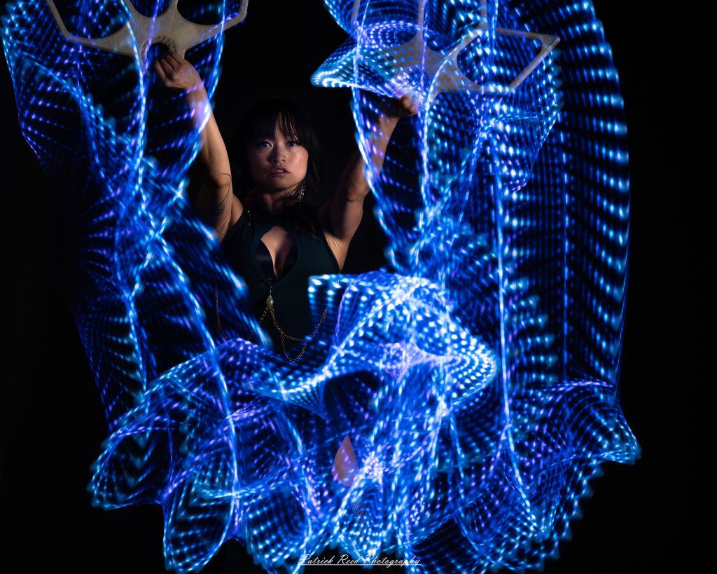 A studio portrait of a model surrounded by flowing light painting patterns created with long-exposure techniques, forming colorful arcs and motion trails against a black background.