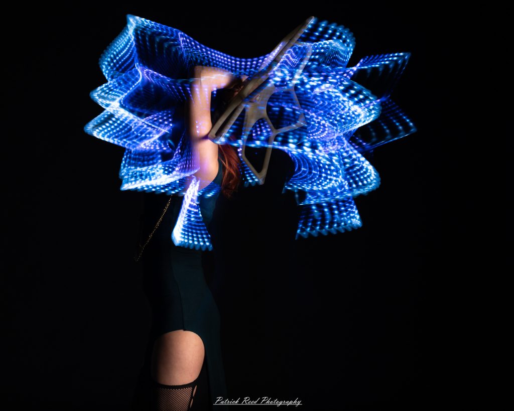 A studio portrait of a model surrounded by flowing light painting patterns created with long-exposure techniques, forming colorful arcs and motion trails against a black background.