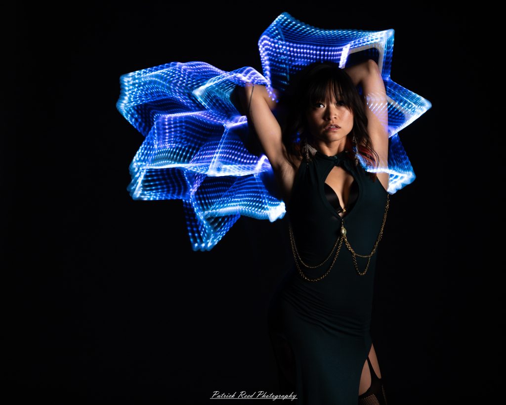 A studio portrait of a model surrounded by flowing light painting patterns created with long-exposure techniques, forming colorful arcs and motion trails against a black background.
