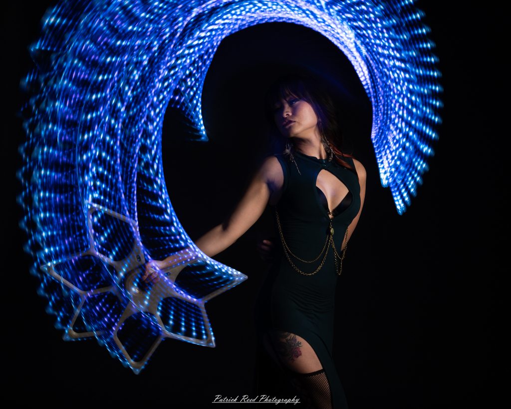 A studio portrait of a model surrounded by flowing light painting patterns created with long-exposure techniques, forming colorful arcs and motion trails against a black background.