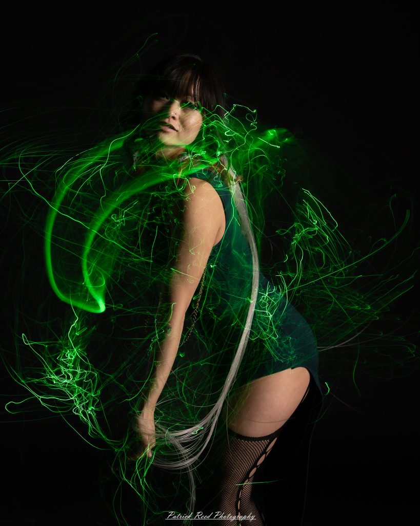 A studio portrait of a model surrounded by flowing light painting patterns created with long-exposure techniques, forming colorful arcs and motion trails against a black background.