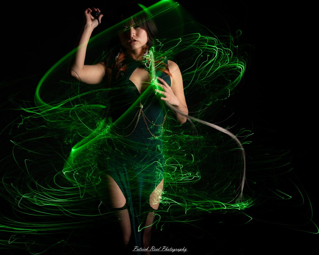 A studio portrait of a model surrounded by flowing light painting patterns created with long-exposure techniques, forming colorful arcs and motion trails against a black background.