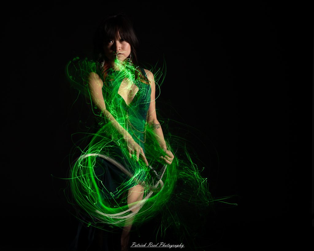 A studio portrait of a model surrounded by flowing light painting patterns created with long-exposure techniques, forming colorful arcs and motion trails against a black background.