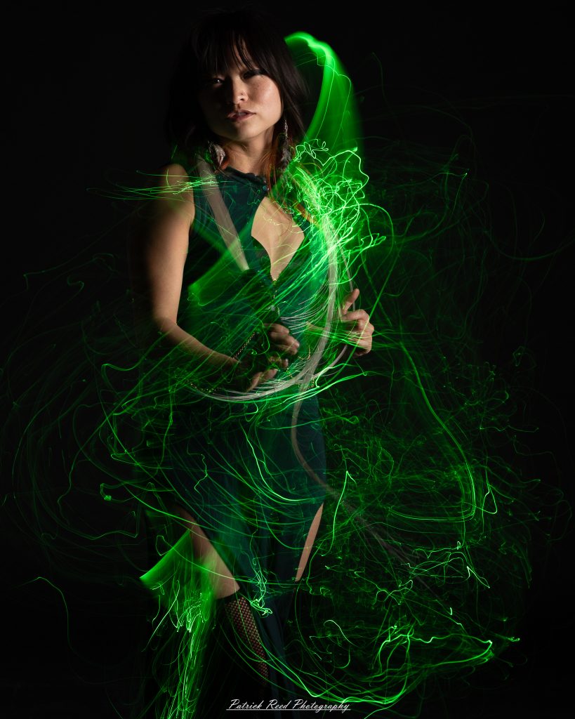 A studio portrait of a model surrounded by flowing light painting patterns created with long-exposure techniques, forming colorful arcs and motion trails against a black background.