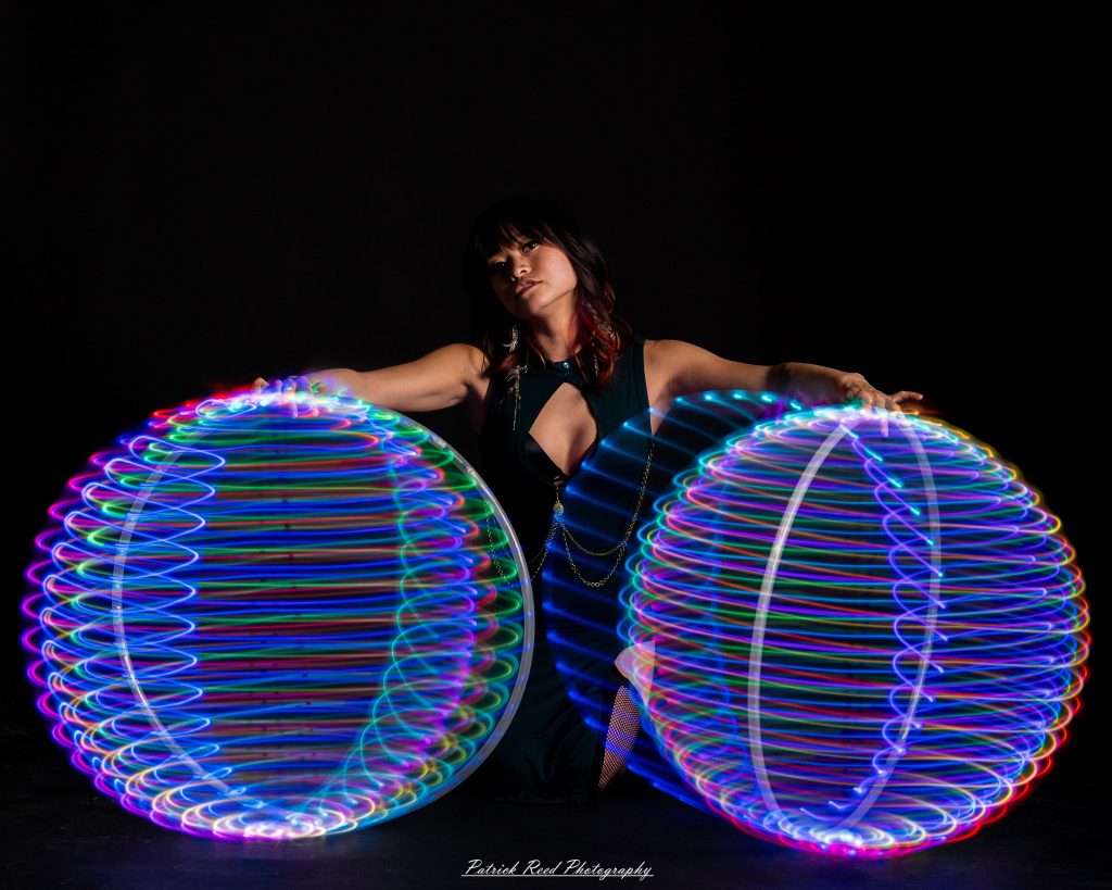 A studio portrait of a model surrounded by flowing light painting patterns created with long-exposure techniques, forming colorful arcs and motion trails against a black background.