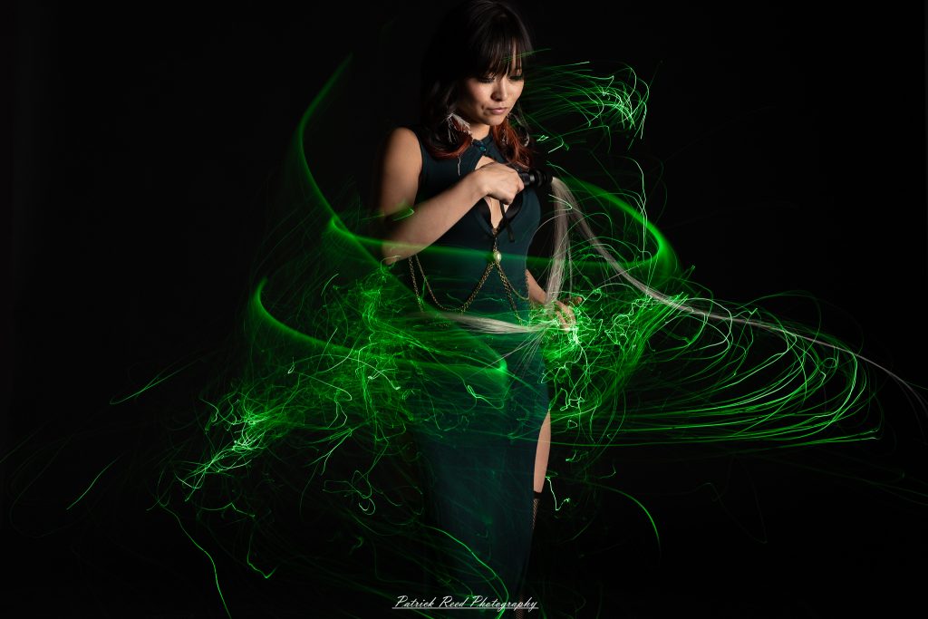 A studio portrait of a model surrounded by flowing light painting patterns created with long-exposure techniques, forming colorful arcs and motion trails against a black background.