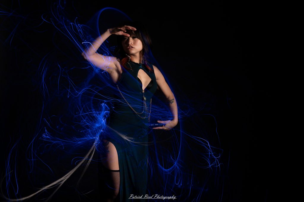 A studio portrait of a model surrounded by flowing light painting patterns created with long-exposure techniques, forming colorful arcs and motion trails against a black background.