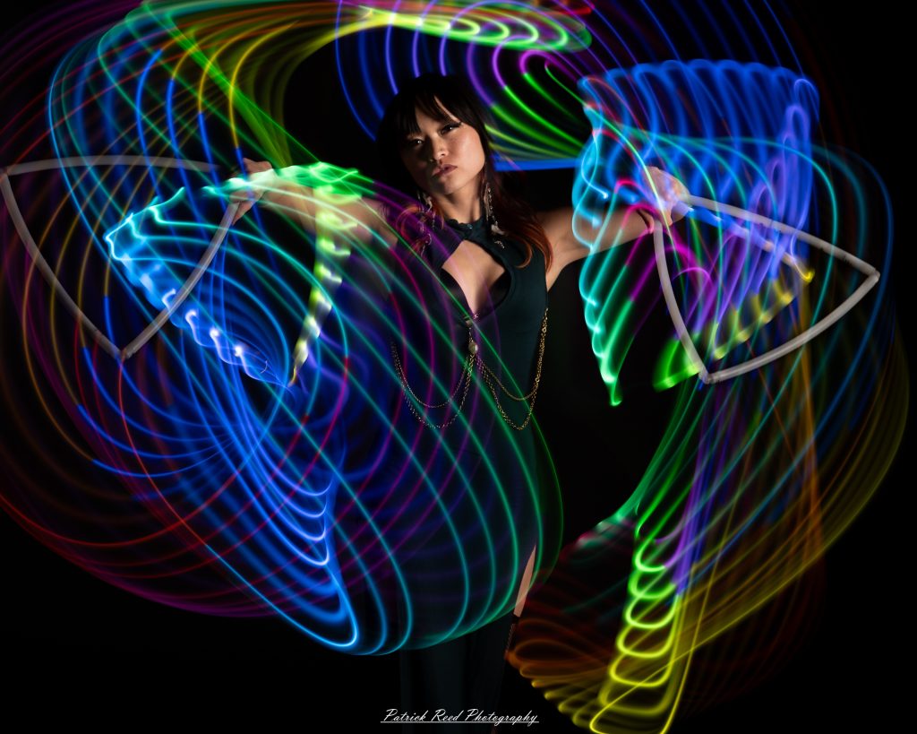 A studio portrait of a model surrounded by flowing light painting patterns created with long-exposure techniques, forming colorful arcs and motion trails against a black background.