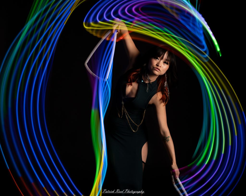 A studio portrait of a model surrounded by flowing light painting patterns created with long-exposure techniques, forming colorful arcs and motion trails against a black background.