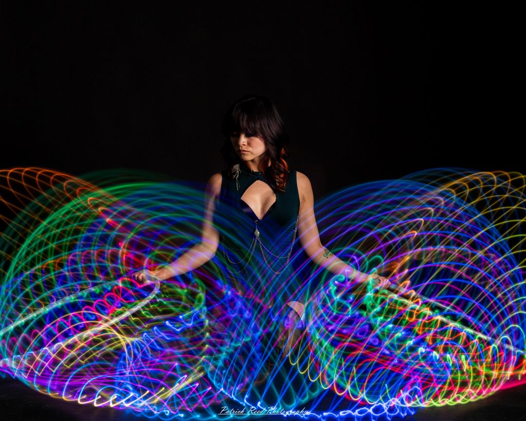 A studio portrait of a model surrounded by flowing light painting patterns created with long-exposure techniques, forming colorful arcs and motion trails against a black background.