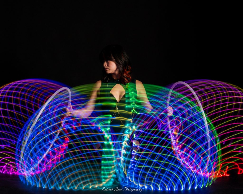 A studio portrait of a model surrounded by flowing light painting patterns created with long-exposure techniques, forming colorful arcs and motion trails against a black background.