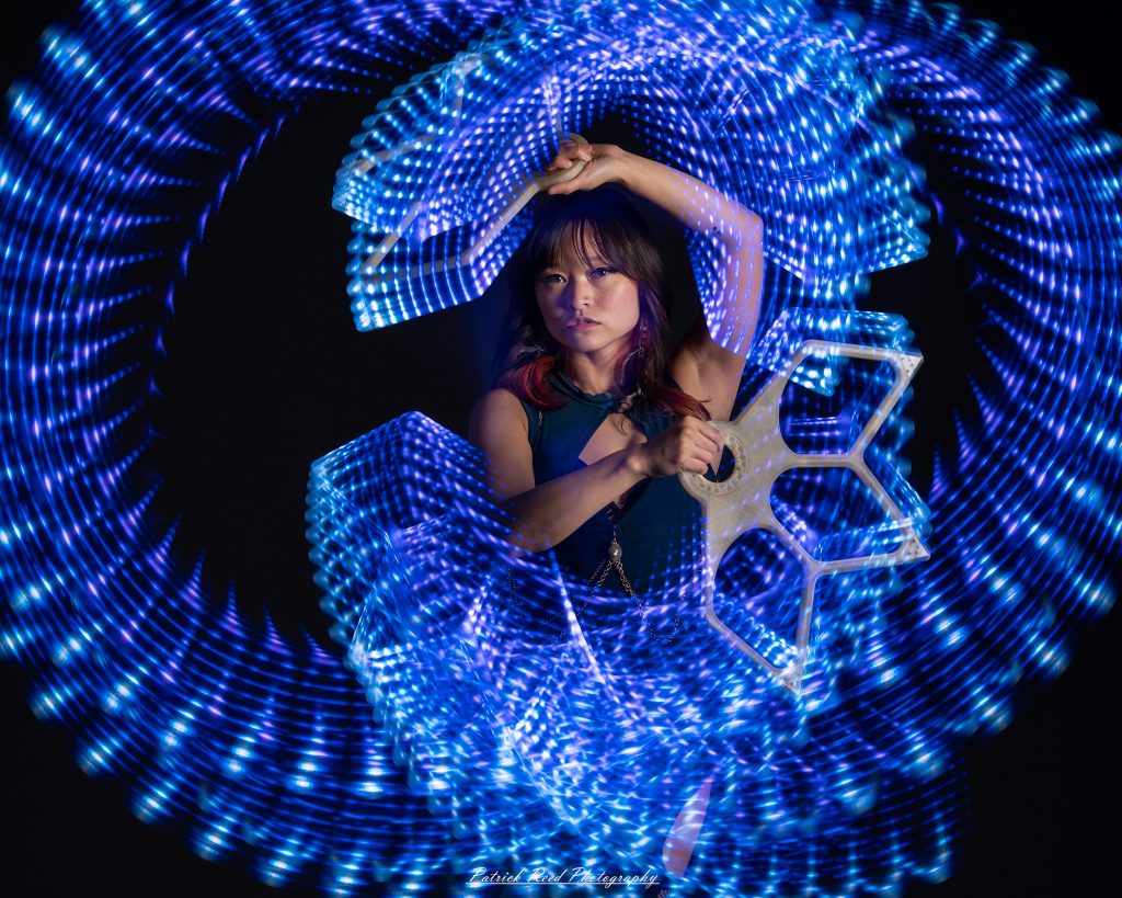 A studio portrait of a model surrounded by flowing light painting patterns created with long-exposure techniques, forming colorful arcs and motion trails against a black background.