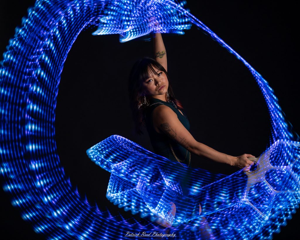 A studio portrait of a model surrounded by flowing light painting patterns created with long-exposure techniques, forming colorful arcs and motion trails against a black background.