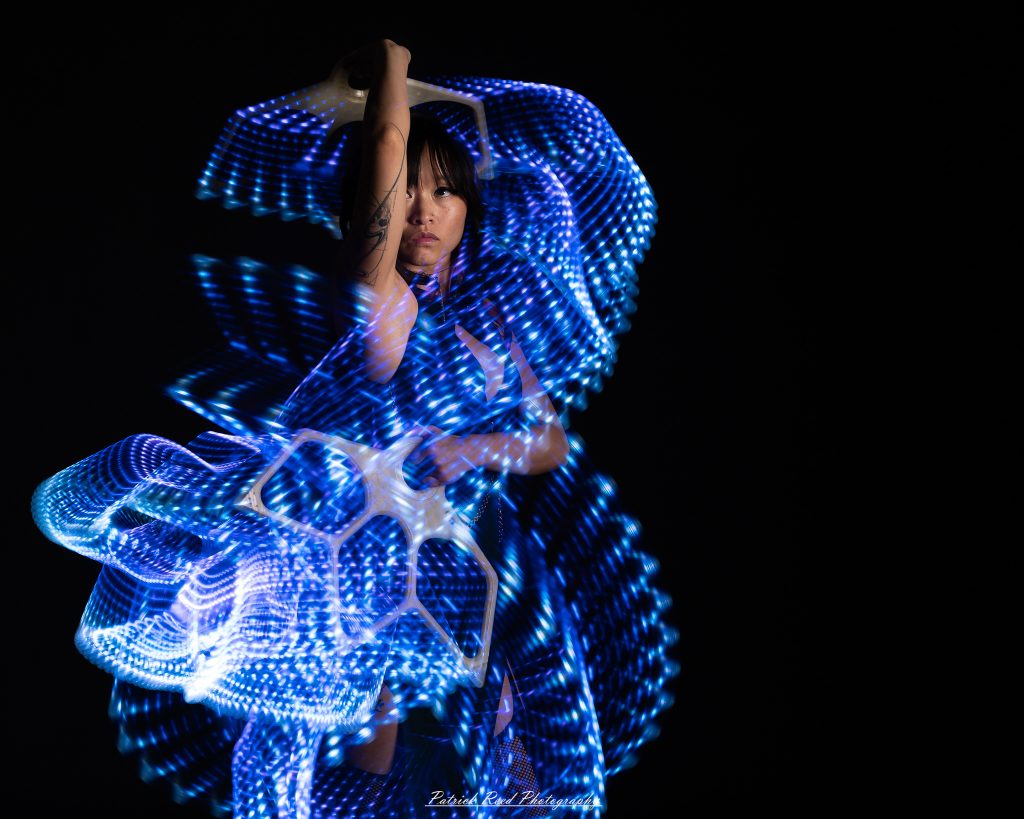 A studio portrait of a model surrounded by flowing light painting patterns created with long-exposure techniques, forming colorful arcs and motion trails against a black background.