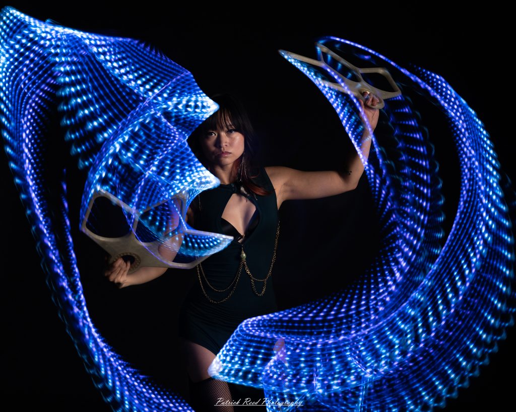 A studio portrait of a model surrounded by flowing light painting patterns created with long-exposure techniques, forming colorful arcs and motion trails against a black background.