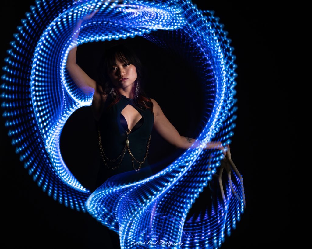 A studio portrait of a model surrounded by flowing light painting patterns created with long-exposure techniques, forming colorful arcs and motion trails against a black background.