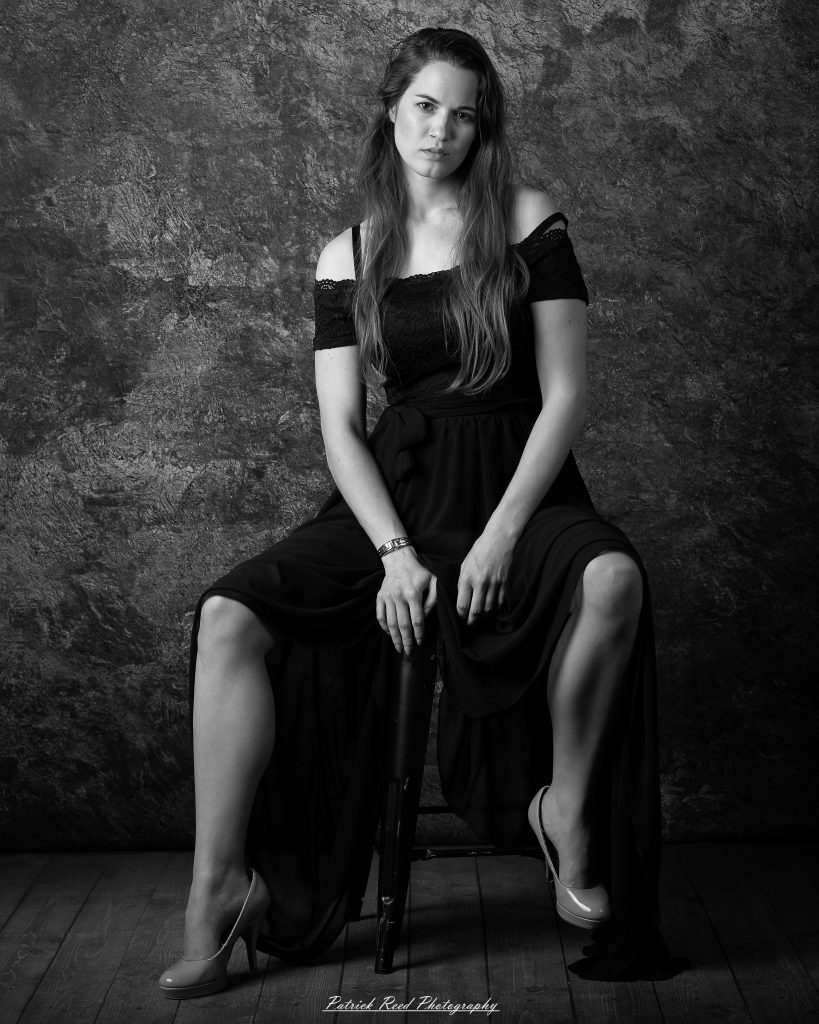 Black-and-white portrait of a woman seated on a stool in a long dark dress against a textured studio backdrop.