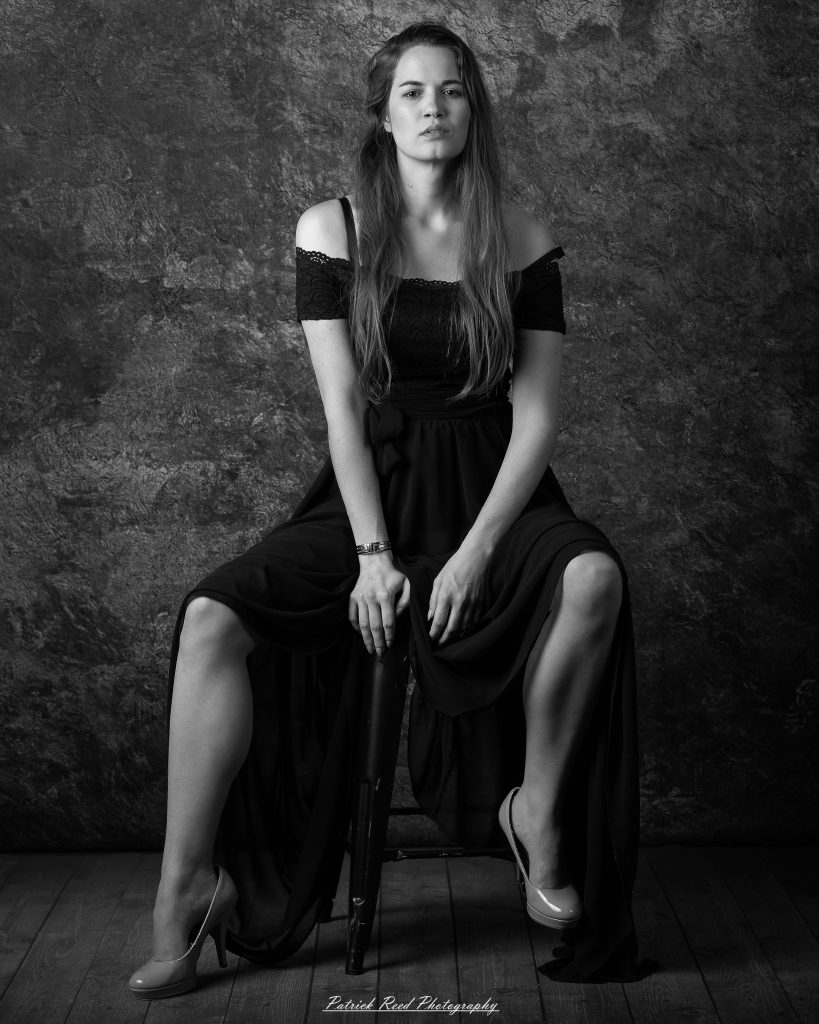 Monochrome portrait of a woman sitting forward with hands resting on a stool, wearing a flowing dark dress.