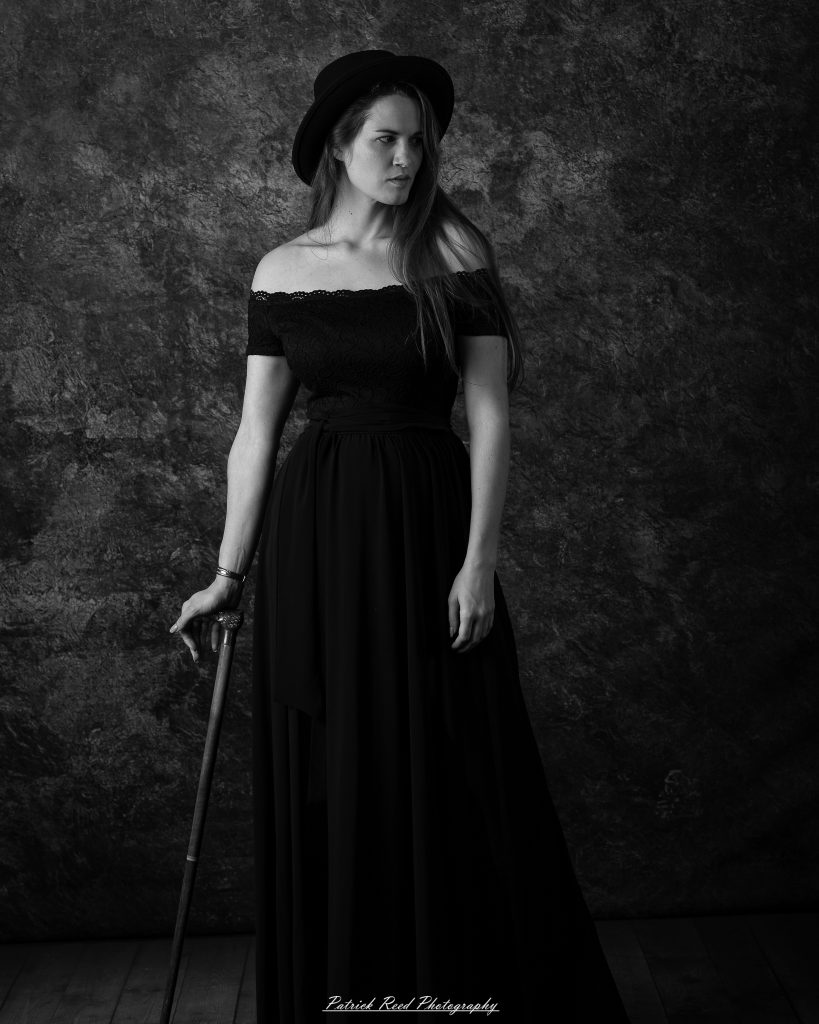 Full-body monochrome portrait of a woman in a long dress and hat, holding a cane in a studio setting.