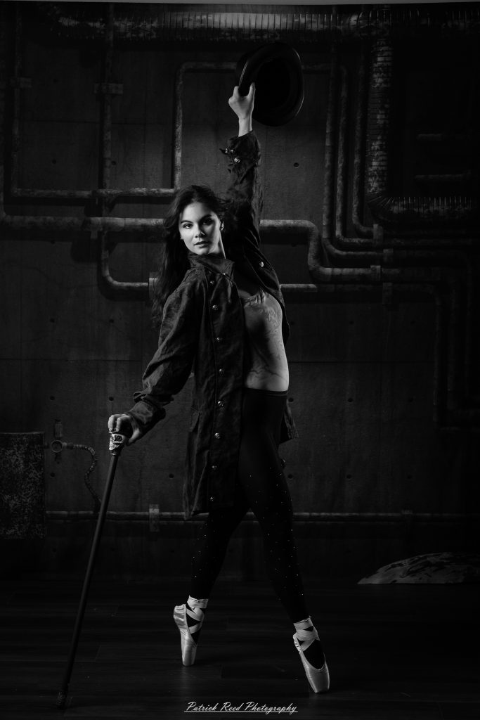 Black and white portrait of a steampunk-inspired dancer wearing a coat, hat, and holding a cane, performing expressive movements in an industrial studio setting with pipes and dramatic lighting.