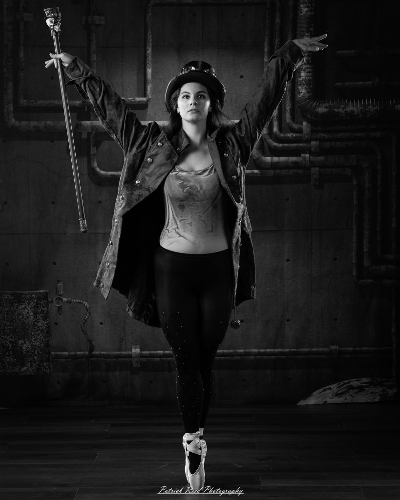 Black and white portrait of a steampunk-inspired dancer wearing a coat, hat, and holding a cane, performing expressive movements in an industrial studio setting with pipes and dramatic lighting.