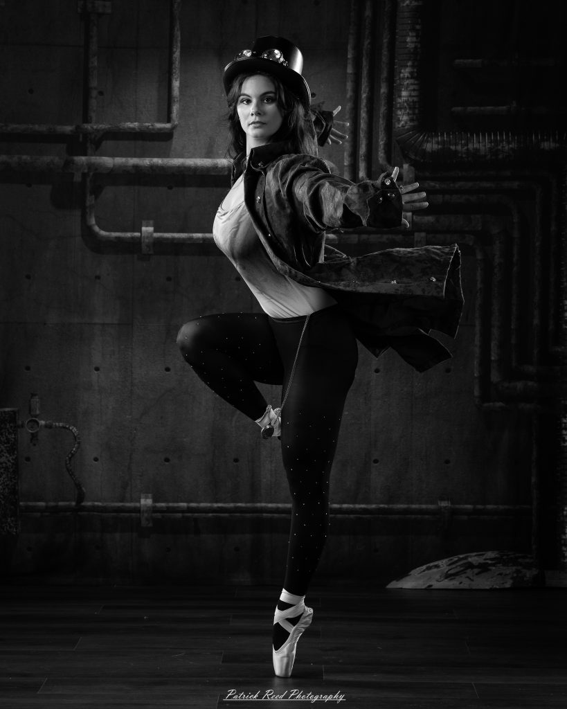 Black and white portrait of a steampunk-inspired dancer wearing a coat, hat, and holding a cane, performing expressive movements in an industrial studio setting with pipes and dramatic lighting.