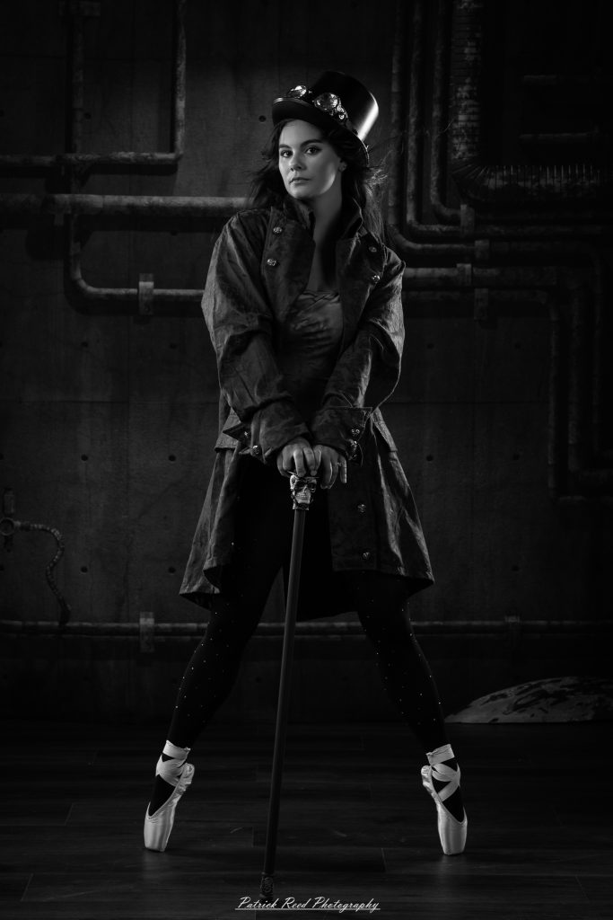 Black and white portrait of a steampunk-inspired dancer wearing a coat, hat, and holding a cane, performing expressive movements in an industrial studio setting with pipes and dramatic lighting.