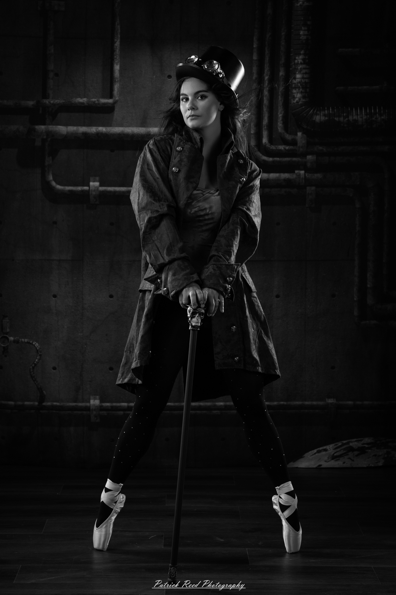 Black and white portrait of a steampunk-inspired dancer wearing a coat, hat, and holding a cane, performing expressive movements in an industrial studio setting with pipes and dramatic lighting.