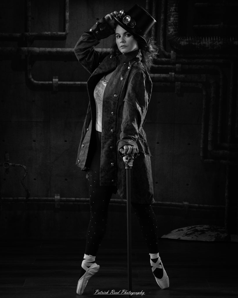 Black and white portrait of a steampunk-inspired dancer wearing a coat, hat, and holding a cane, performing expressive movements in an industrial studio setting with pipes and dramatic lighting.