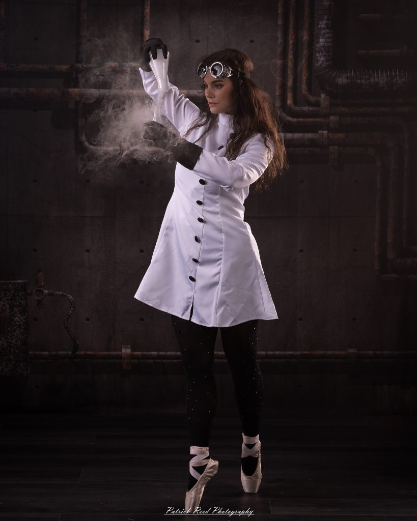 Black and white portrait of a dancer in a white coat performing a mad scientist concept with powder and smoke effects, captured in motion against an industrial studio backdrop.
