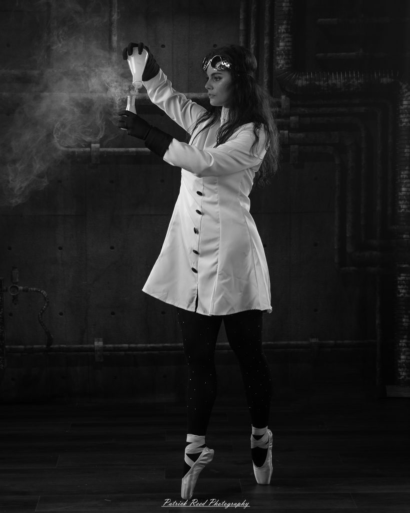 Black and white portrait of a dancer in a white coat performing a mad scientist concept with powder and smoke effects, captured in motion against an industrial studio backdrop.