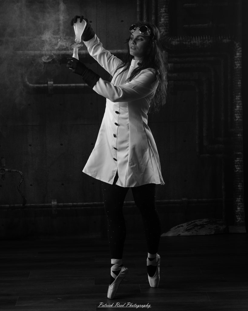Black and white portrait of a dancer in a white coat performing a mad scientist concept with powder and smoke effects, captured in motion against an industrial studio backdrop.