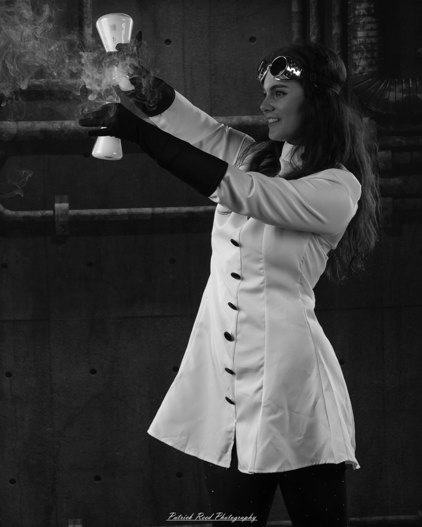 Black and white portrait of a dancer in a white coat performing a mad scientist concept with powder and smoke effects, captured in motion against an industrial studio backdrop.