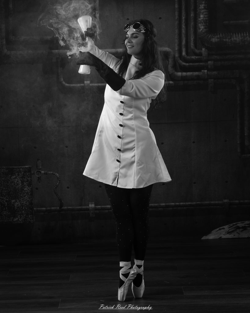 Black and white portrait of a dancer in a white coat performing a mad scientist concept with powder and smoke effects, captured in motion against an industrial studio backdrop.