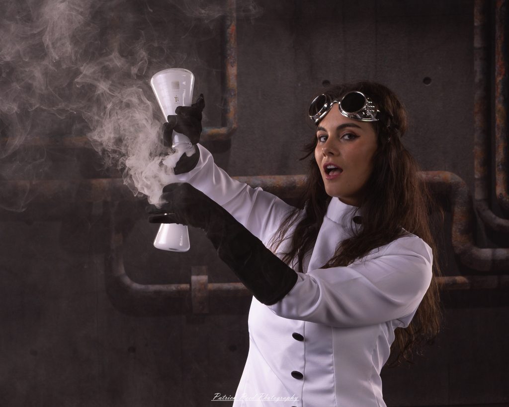 Black and white portrait of a dancer in a white coat performing a mad scientist concept with powder and smoke effects, captured in motion against an industrial studio backdrop.
