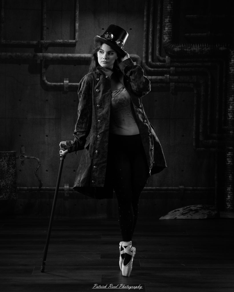 Black and white portrait of a steampunk-inspired dancer wearing a coat, hat, and holding a cane, performing expressive movements in an industrial studio setting with pipes and dramatic lighting.