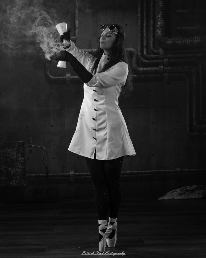 Black and white portrait of a dancer in a white coat performing a mad scientist concept with powder and smoke effects, captured in motion against an industrial studio backdrop.