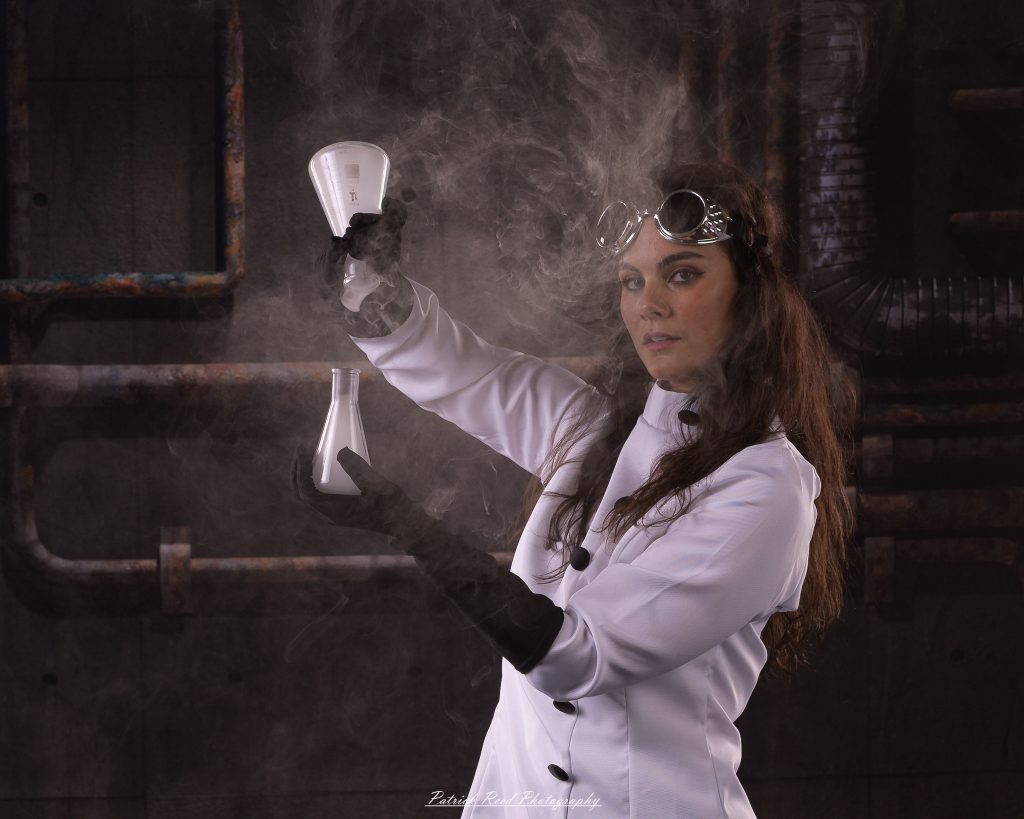 Black and white portrait of a dancer in a white coat performing a mad scientist concept with powder and smoke effects, captured in motion against an industrial studio backdrop.