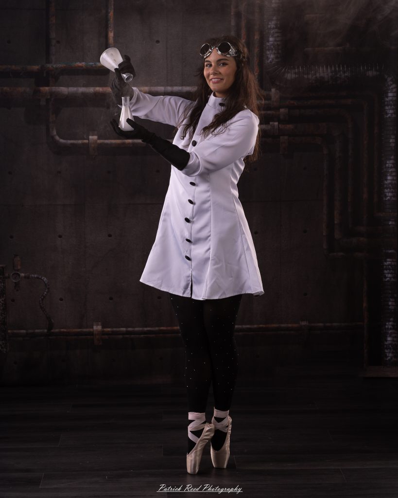 Black and white portrait of a dancer in a white coat performing a mad scientist concept with powder and smoke effects, captured in motion against an industrial studio backdrop.