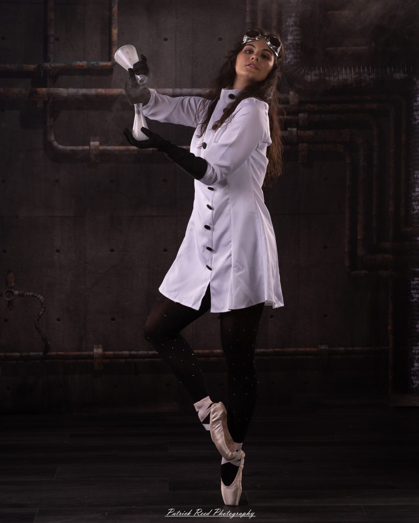 Black and white portrait of a dancer in a white coat performing a mad scientist concept with powder and smoke effects, captured in motion against an industrial studio backdrop.