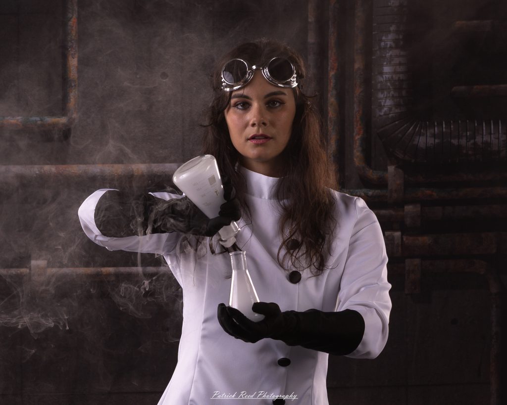 Black and white portrait of a dancer in a white coat performing a mad scientist concept with powder and smoke effects, captured in motion against an industrial studio backdrop.