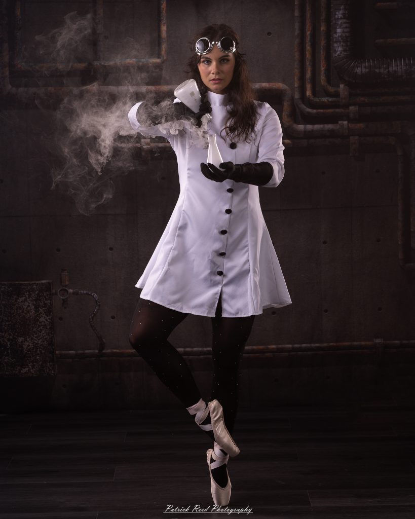 Black and white portrait of a dancer in a white coat performing a mad scientist concept with powder and smoke effects, captured in motion against an industrial studio backdrop.