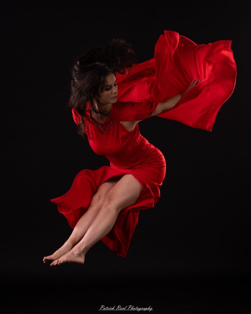 Black and white fine art dance portrait of a dancer in a flowing dress, captured in motion with expressive movement and soft studio lighting against a dark background.