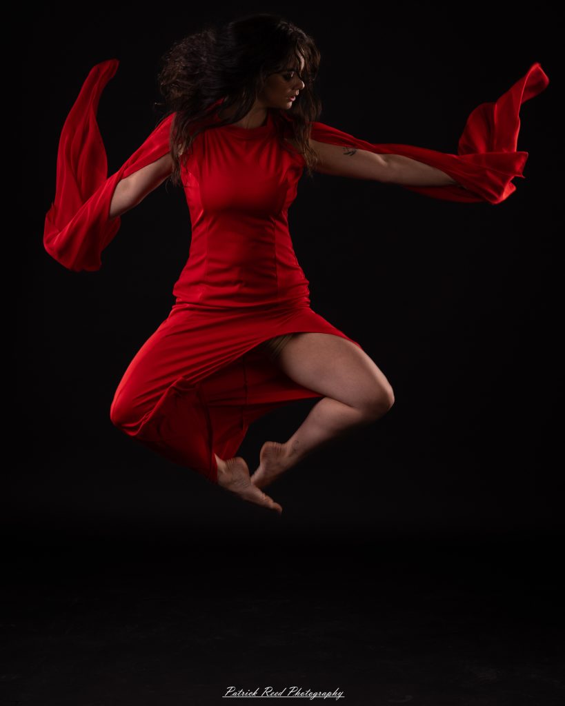 Black and white fine art dance portrait of a dancer in a flowing dress, captured in motion with expressive movement and soft studio lighting against a dark background.
