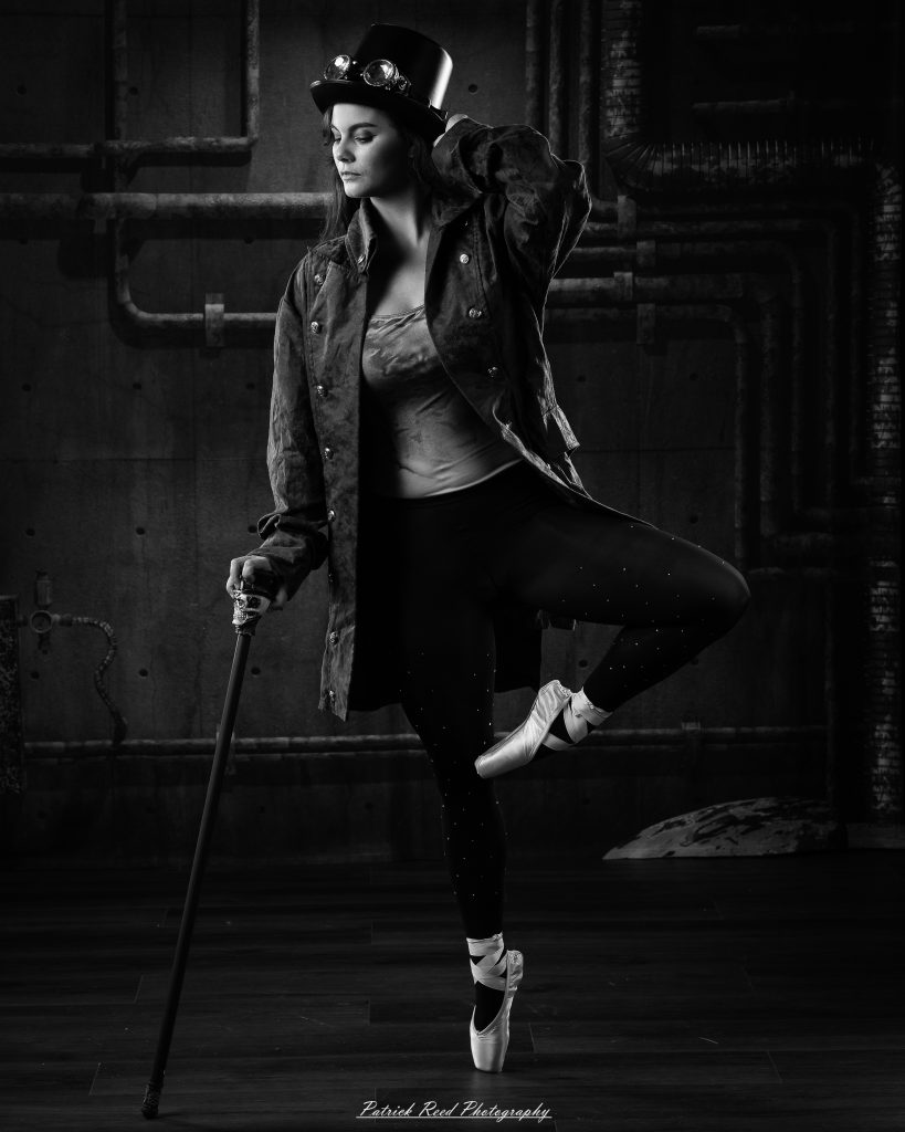 Black and white portrait of a steampunk-inspired dancer wearing a coat, hat, and holding a cane, performing expressive movements in an industrial studio setting with pipes and dramatic lighting.