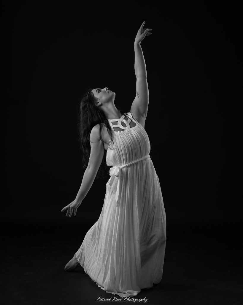 Black and white fine art dance portrait of a dancer in a flowing dress, captured in motion with expressive movement and soft studio lighting against a dark background.