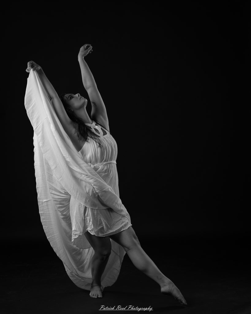 Black and white fine art dance portrait of a dancer in a flowing dress, captured in motion with expressive movement and soft studio lighting against a dark background.