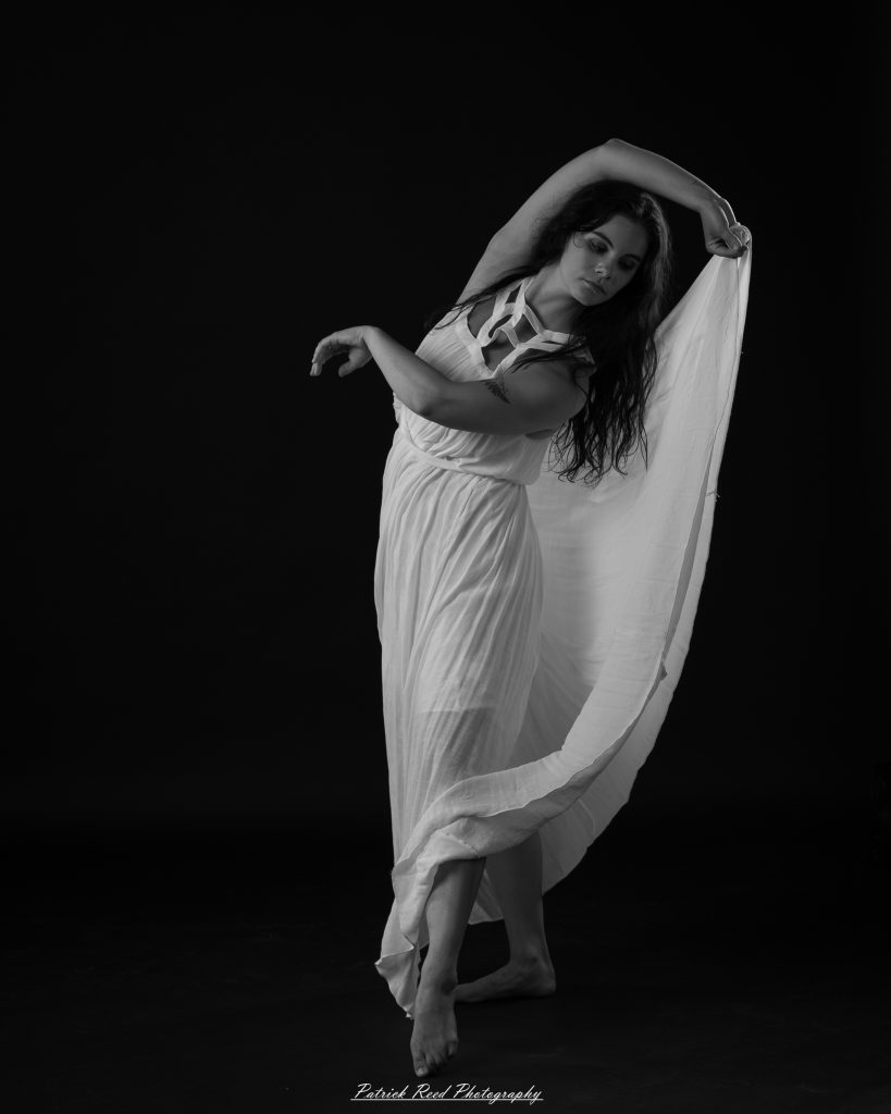 Black and white fine art dance portrait of a dancer in a flowing dress, captured in motion with expressive movement and soft studio lighting against a dark background.