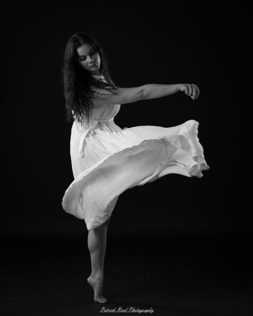 Black and white fine art dance portrait of a dancer in a flowing dress, captured in motion with expressive movement and soft studio lighting against a dark background.