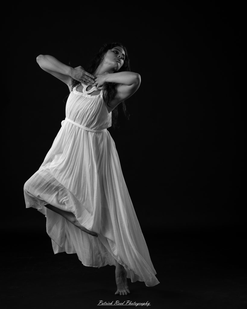 Black and white fine art dance portrait of a dancer in a flowing dress, captured in motion with expressive movement and soft studio lighting against a dark background.